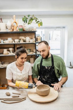 Smiling female artisan in apron drying ceramic bowl with heat gun near boyfriend in ceramic workshop clipart