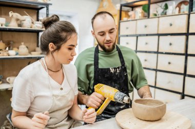 Craftsman in apron drying ceramic bowl with heat gun on wooden board near girlfriend in studio clipart