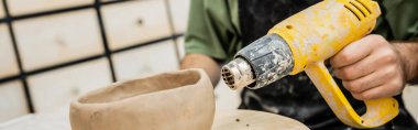 Cropped view of potter drying clay bowl with heat gun on wooden board in pottery workshop, banner clipart