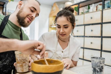 Smiling couple of sculptors in aprons talking and coloring clay bowl in ceramic studio at background clipart