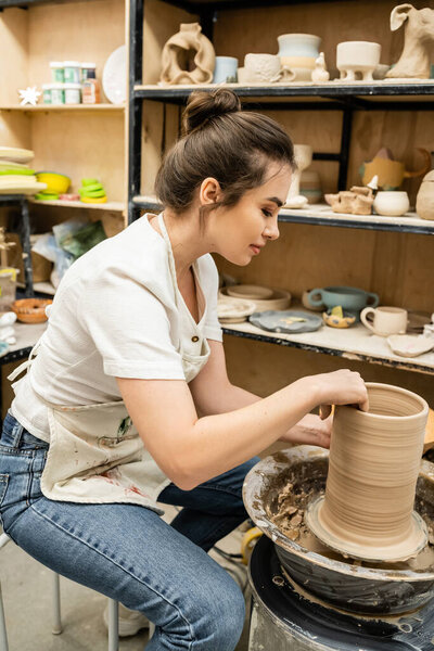 Side view of brunette craftswoman in apron working with clay on pottery wheel in studio in workshop