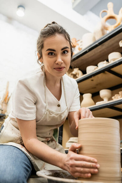 Brunette craftswoman in apron looking at camera and making clay vase on pottery wheel in workshop