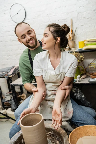 Stock image Positive artisan hugging girlfriend in apron and making clay vase on pottery wheel in studio