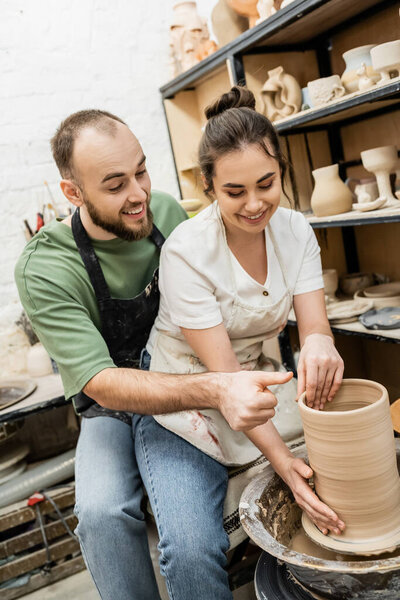Positive sculptor talking to girlfriend making clay vase on pottery wheel in ceramic workshop