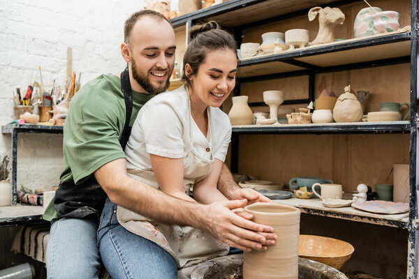 Smiling romantic couple in aprons shaping clay vase on pottery wheel in ceramic workshop at background