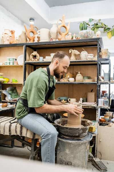 Side view of potter in apron shaping clay sculpture on pottery wheel near rack in ceramic workshop