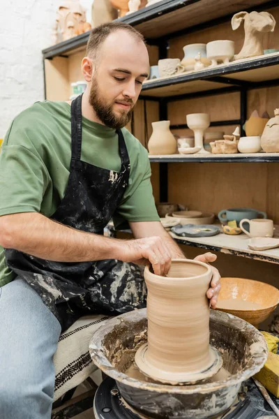 stock image Bearded craftsman in apron making clay vase on pottery wheel near rack with sculptures in workshop