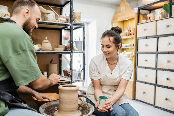 Smiling craftswoman holding cup while boyfriend making clay vase on pottery wheel in studio