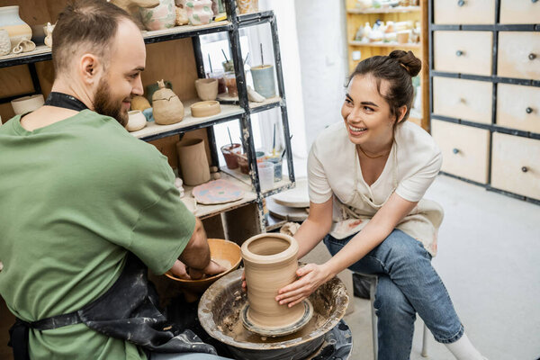 Smiling craftswoman shaping clay vase on pottery wheel near boyfriend and bowl with water in studio