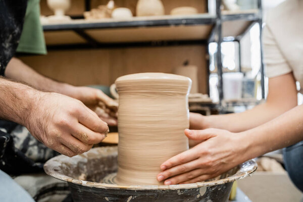 Cropped view of romantic artisans shaping clay vase on pottery wheel together in ceramic studio