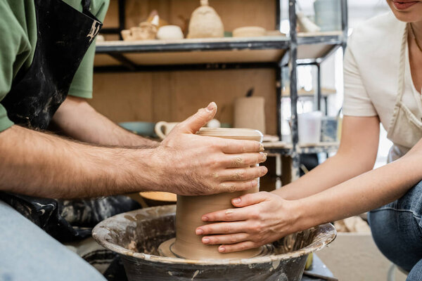 Cropped view of couple of artisans making clay vase on pottery wheel near rack in ceramic workshop