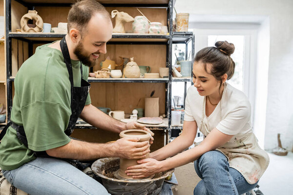 Positive couple of potters shaping clay vase on pottery wheel together in ceramic studio