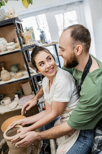 Joyful craftswoman in apron looking at boyfriend and molding clay on pottery wheel in workshop
