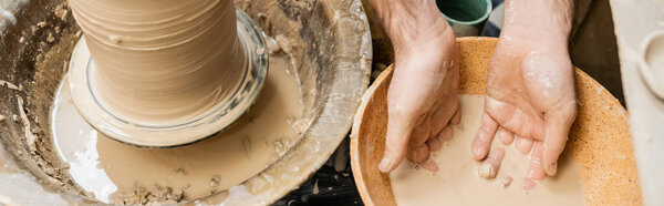 Top view of artisan working with water in bowl near clay and pottery wheel in ceramic studio, banner