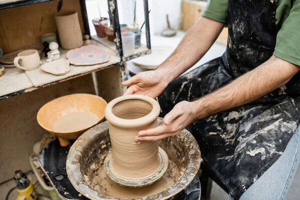 Cropped view of potter in dirty apron shaping clay vase on pottery wheel near rack in workshop