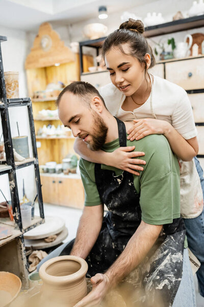 Smiling female potter in apron hugging boyfriend working with clay and pottery wheel in workshop