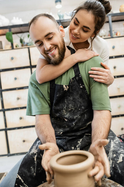 Smiling craftswoman embracing boyfriend in apron shaping clay vase on pottery wheel in studio