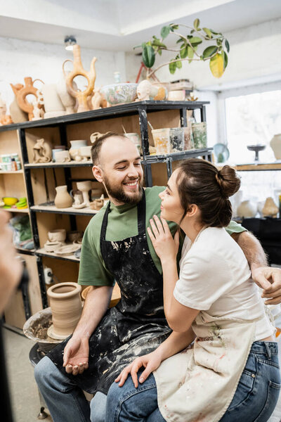 Joyful bearded sculptor in apron hugging girlfriend near blurred clay and pottery wheel in workshop