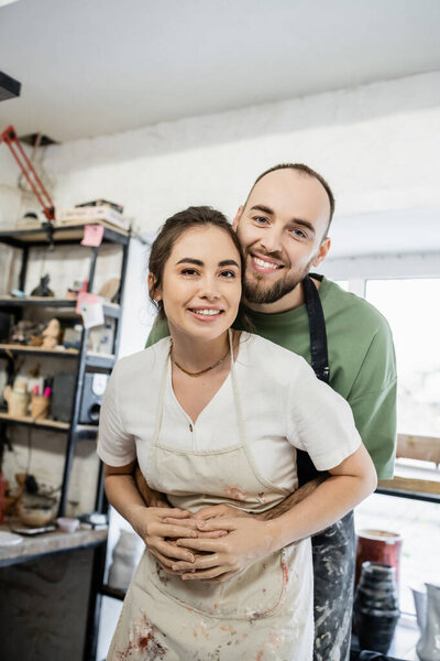 Positive bearded craftsman hugging girlfriend in apron and looking at camera in ceramic workshop
