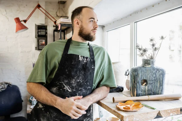 stock image Bearded craftsman in apron looking away while standing near vase with flowers in ceramic workshop