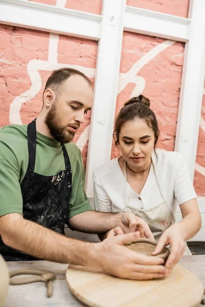 stock image Bearded craftsman in apron shaping clay bowl and talking to girlfriend in ceramic workshop