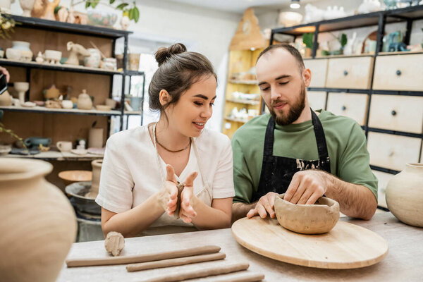 Smiling craftswoman in apron molding clay while boyfriend making bowl in ceramic workshop