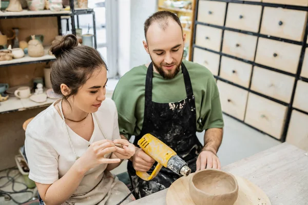 stock image Couple of artisans in aprons working and drying clay with heat gun while talking in workshop