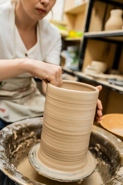 Cropped view of female artisan in apron shaping clay on pottery wheel in blurred workshop