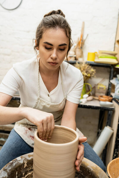 Ремесленница брюнеток в фартуке, облицовывающая глину на гончарном круге керамической студии на заднем плане