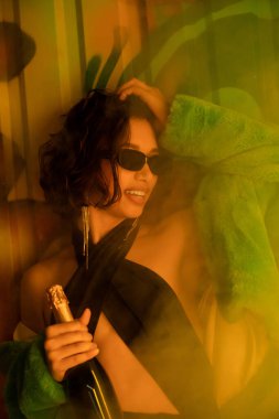 Stylish cheerful woman in sunglasses holding champagne near graffiti and smoke in night club clipart
