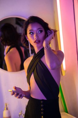 Gece kulübünde elinde akıllı telefon tutan ve saçlarına neon ışıkta dokunan modaya uyan Asyalı genç bir kadın.