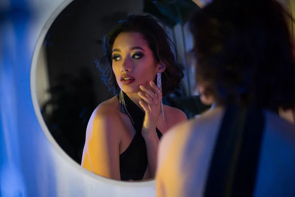 stock image Fashionable young asian woman in dress looking away near mirror in neon light in night club