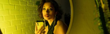 Asyalı trend bir kadın akıllı telefonuyla selfie çekiyor. Aynanın yanında, gece kulübünde, pankartta.