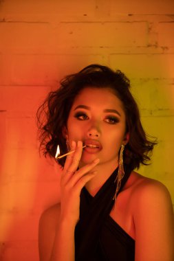 Fashionable young asian woman holding match with fire in neon light in night club clipart