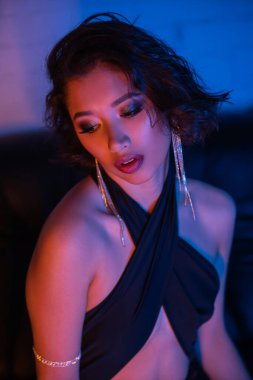 Saç stili ve makyajlı seksi Asyalı kadın gece kulübünde neon ışıklı koltukta oturuyor.