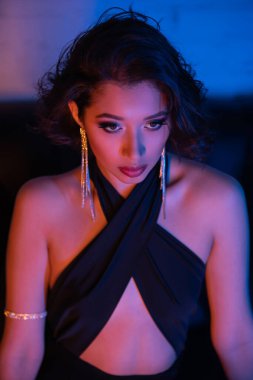 Şık elbiseli ve küpeli kendine güvenen Asyalı kadın gece kulübünde neon ışıkta kanepede oturuyor.