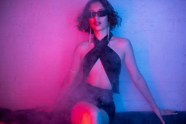 Seksi elbiseli ve güneş gözlüklü şık Asyalı kadın gece kulübünde duman ve neon ışıkta poz veriyor.