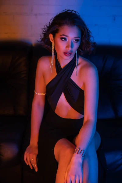 Trendy asian woman in earrings, bracelets and dress sitting on couch with neon light in night club