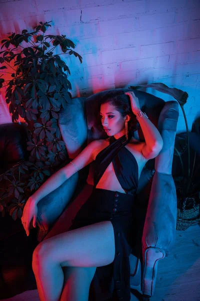 Well-dressed asian woman in dress sitting in armchair near plants in night club with neon light