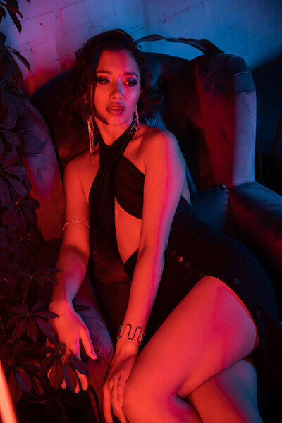 Sexy and trendy asian woman sitting in armchair near plants in colorful neon light in night club