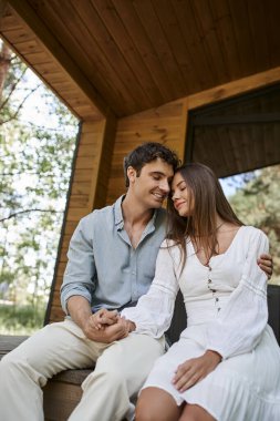 Romantik bir adam verandada oturup, el ele tutuşurken, neşeli bir kadına sarılıyor.