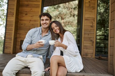 Kahvelerini yudumlayan neşeli bir çift, bir adam ve bir kadın tatil evinin verandasında gülüyor, sabah