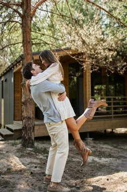 happy man lifting and hugging woman in vacation house, forest, look at each other, romance and love clipart
