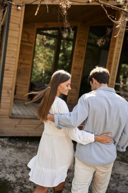 Smiling brunette woman in sundress hugging boyfriend and walking near summer house outdoors clipart