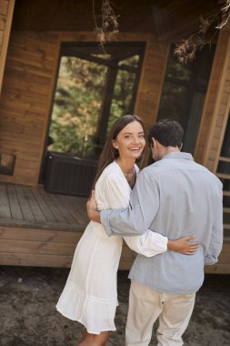 Joyful young woman in sundress hugging boyfriend and looking at camera near vacation house outdoors clipart