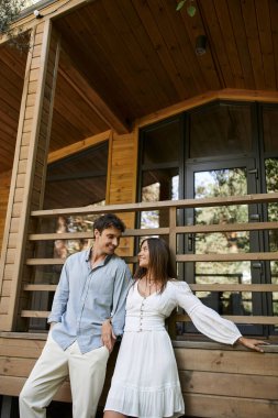 Gülümseyen romantik çift, modaya uygun giyinmiş arka planda tatil evinin yanında duruyor.