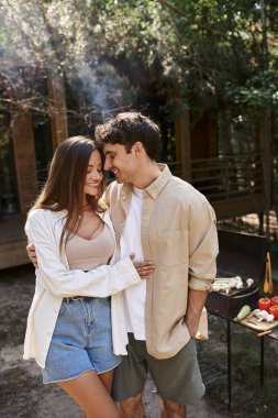 Gülümseyen romantik çift arka planda bulanık barbekü ve tatil evinin yakınında sarılıyorlar.