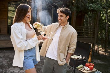 Kız arkadaşının yanına şarap döken gülümseyen adam, arka planda bulanık barbekü ve tatil evi.