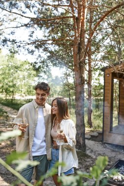 Neşeli bir çift gündelik giysiler içinde ellerinde şarap ve arka planda bulanık tatil evi tutuyorlar.