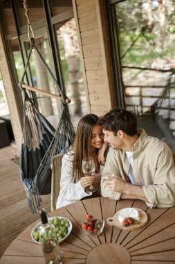 Positive woman hugging boyfriend near wine and food on terrace of wooden summer house clipart